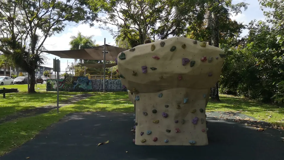 Sand-coloured artificial boulder with coloured holds on black rubber matting, with the skate park's shade sails behind