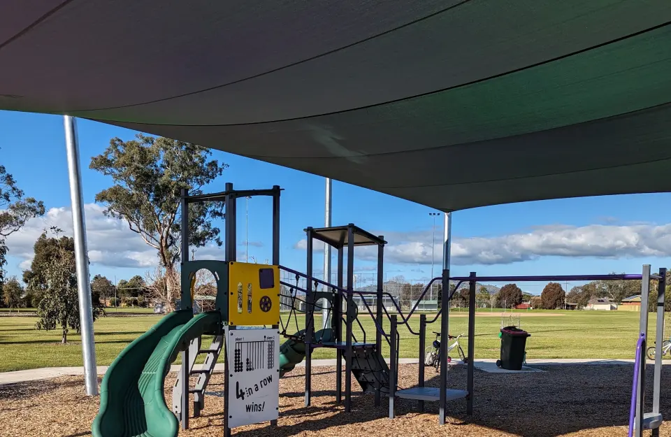 Play unit with green slides under a shade sail on the bark-chip softfall, with Gayview Park's grassy field behind
