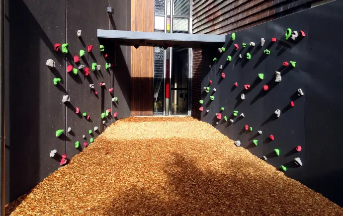 Black bouldering panels with green, red and grey holds above a bed of bark chips