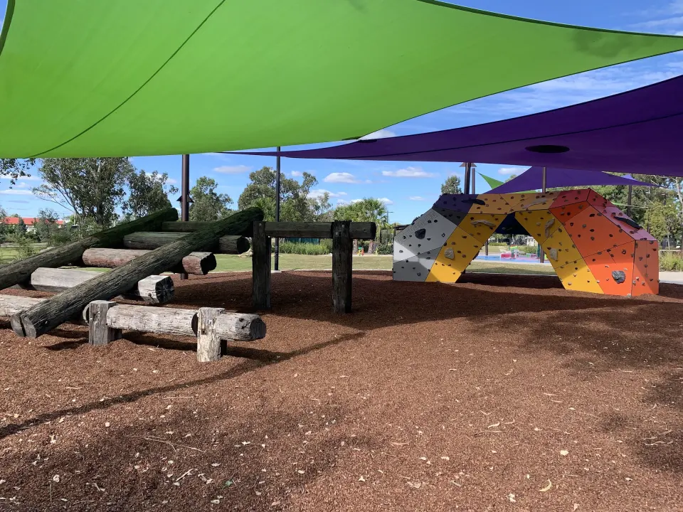 Nature-play logs on bark softfall, with the bouldering arch and the water park beyond